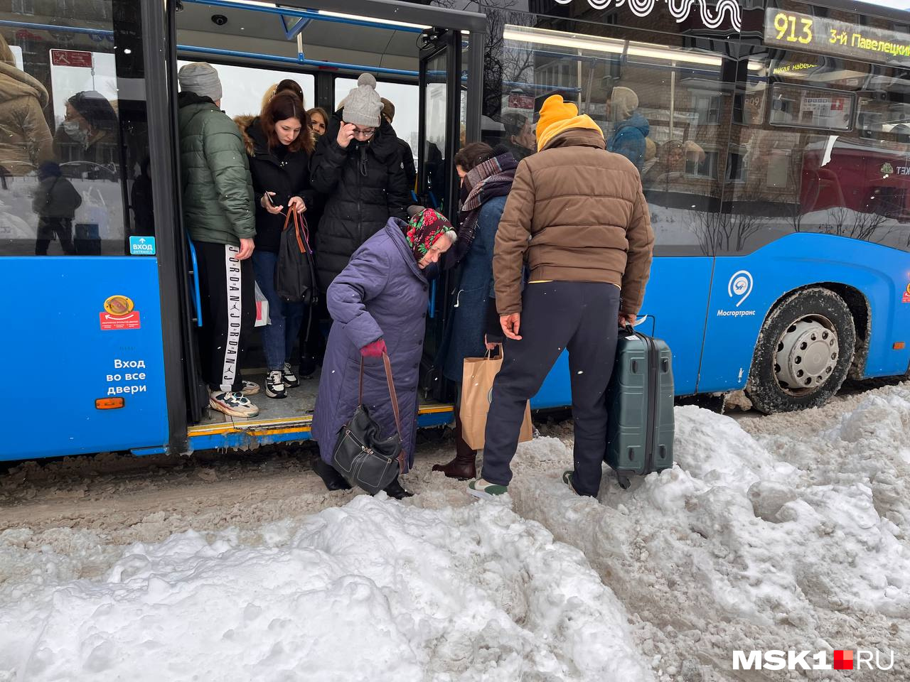 Пожилая женщина с трудом выходит из автобуса, который и без того выше уровня земли, и наступает прямо в сугроб