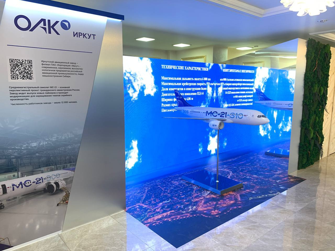 Макет самолета МС-21, который собирают на Иркутском авиазаводе, на фоне экрана с облаками и информацией о производстве корпорации «Иркут»