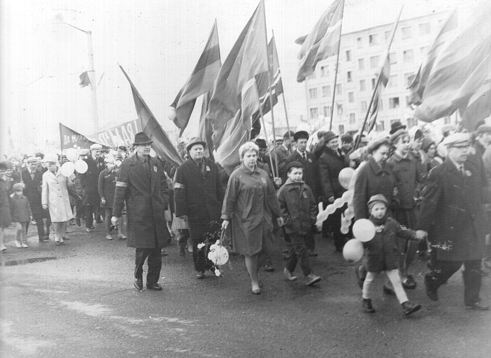 Горожане на праздничной демонстрации. В руках люди несут флаги, ветки и шарики. Дети участвуют в шествии в сопровождении взрослых